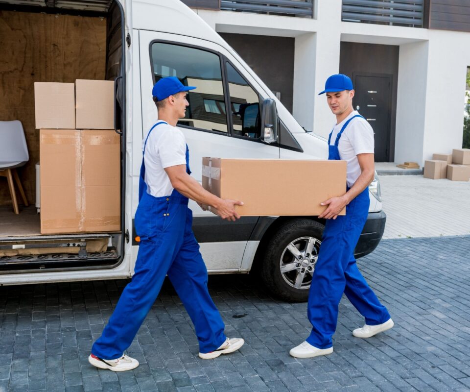 Two movers in blue overalls carry a large box from a white delivery van beside a modern house behind them.